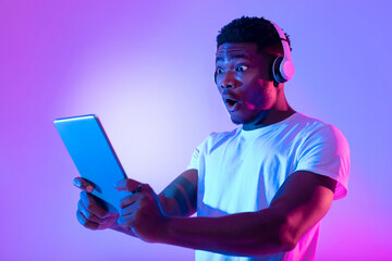 Poster - Shocked black guy in headphones looking at tablet with glowing screen, opening mouth in surprise, neon lighting