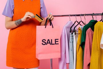 cropped view of tattooed woman with credit card and sale shopping bag near rack with garments isolated on pink.