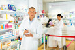 © JackF - Doctor in white coat standing in salesroom with pan and recipe blanks in hands and looking in camera. His co-worker setting out drugs on shelves in background.