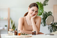 Beautiful Smiling Woman Sitting At Table With Bottles Of Essential Oil In Room
