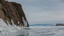 A Sheer Granite Rock, Devoid Of Vegetation, Rises On A Frozen Lake. The Base Is Icy. A Mountain Range Against A Cloudy Sky. Reflection On The Ice. Baikal