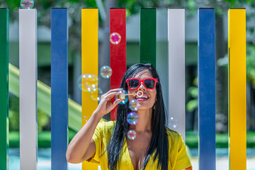 Latina woman smiling and blowing bubbles soap in a yellow dress against a background of a pool and colorful bars on a sunny day