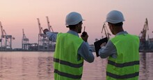 Men In Helmets And Reflective Vests Stand By Seaside And Regulate Work Of Portal Cranes Via Tablet And Walkie-talkie During Sunset