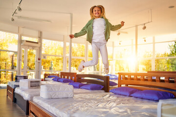 Wall Mural - Beautiful child cute girl jumping on the bed and having fun in the store. Purchase of bedding, mattress and bed.