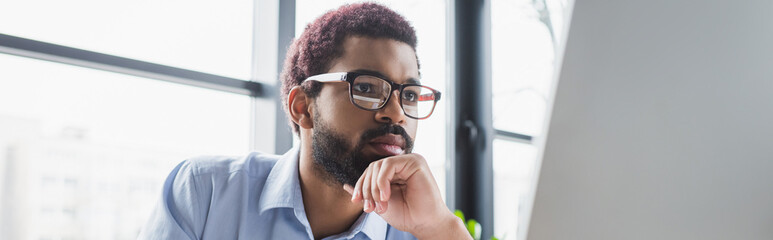 Focused african american businessman in eyeglasses looking at computer monitor in office, banner