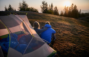 Wall Mural - Back view of two travelers man and woman sitting on grass in front of tent set up on mountain hill, early in the morning and contemplating on sunny sky. Concept of hiking, camping and relationships.