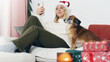 © artshake - Young smiling woman wearing a santa hat making video call on cosy couch with puppy dog christmas gifts and decoration around