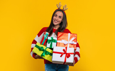 Wall Mural - Cute smiling excited young brunette woman in deer hat, with many colorful Christmas gift boxes in hands isolated on yellow background.