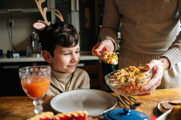 Wall Mural - Cropped shot of unrecognizable young male father putting salad on plate to loving child son, preparing Christmas table at home for family party. Man serving beautiful table for friendly feast.