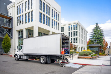 Day cab big rig white semi truck with open long box trailer unloading goods of a moving family standing on the street with multilevel apartments buildings