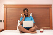 © Confidence - young african woman using her laptop in bed, making a phone call