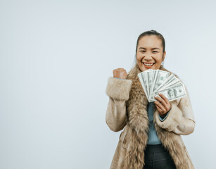 Portrait of successful Asian woman with a fur coat holding money, clenching in fist, smiling and looking to the camera, standing over background in studio with copy space.