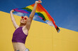 Young non-binary person looking happy and excited while waving a rainbow flag outdoors.