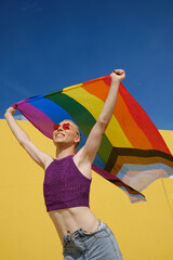 Wall Mural - Low angle view of a young happy non-binary person holding and waving a rainbow flag over head.