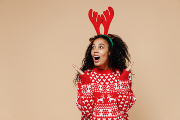 Amazed young african Santa woman in Christmas sweater fun decorative deer horns on head look aside omg isolated on pastel beige background studio portrait. Happy New Year merry x-mas holiday concept.