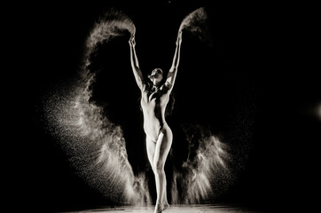 The girl with the flour on the body stretches the arms up with thrown flour on black background black and white image