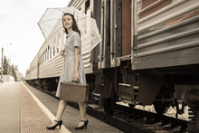 An Elegant Lady In A Dress With An Umbrella And A Vintage Suitcase Gets Off The Train On The Platform. Elegant Lady In Retro Style. A Young Beautiful Woman Arrived By Train.