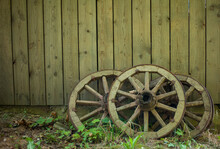 Old Wooden Wagon Wheel Reclining On Wooden Wall. Wheel With Boardwalk Wall On Background And Cobbles On Foreground. Place For Your Text. Wheel Leaning Up Against Wall