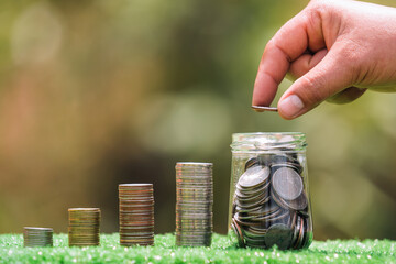 Poster - hand holding coins putting into jug glass with money stack on green grass. concept saving finance and accounting concept