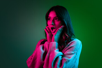 Wall Mural - Studio shot of young adorable cute girl, student in casual style clothes posing isolated on dark green studio backgroud in pink neon light.