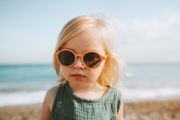 Wall Mural - Child girl traveling walking on beach 3 years old toddler in sunglasses baby portrait looking at camera family vacations trip