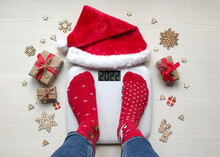 Female Feet Standing On Electronic Scales For Weight Control In Funny Red Christmas Socks On White Wooden Background.