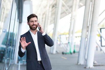 Successful male boss businessman talking outdoors near office uses cellphone confident smiles and rejoices