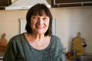 Portrait of happy senior lady sitting in kitchen at home, close-up, copy space. Attractive old brunette woman with short hair smiling at camera resting after preparing dinner for her family