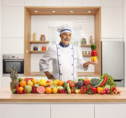 Poster - Mature male chef holding a pile of bell peppers and standing behind fruits and vegetables