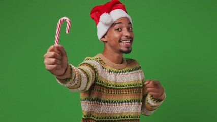 Wall Mural - Merry fun young african american Santa man 20s wears striped shirt Christmas hat pointing finger on candy cane show thumb up like gesture isolated on plain olive green khaki background studio portrait