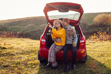 Loving couple relaxing in car trunk by autumn lake drinking tea from vacuum flask. Man and woman travel by auto.