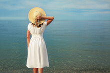 A Young Girl In A White Dress Stands On The Seashore, Looking Into The Distance. Holds The Straw Hat With His Right Hand