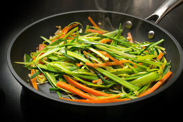 Steaming vegetable noodles from fresh carrot julienne and green leek strips in a black frying pan, cooking a healthy vegetarian meal, selected focus