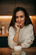 © Александра Шамонина - A young brunette girl with long wavy hair stands in the kitchen at home in a white sweater with a mug of coffee, looks into the frame