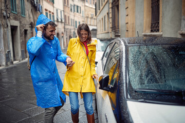 Wall Mural - A young cheerful couple in raincoats reached the car while running away from the rain in the city. Walk, rain, city, relationship