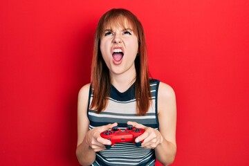 Redhead young woman playing video game holding controller angry and mad screaming frustrated and furious, shouting with anger looking up.