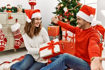 Wall Mural - Young hispanic couple wearing christmas hat opening gift at home.