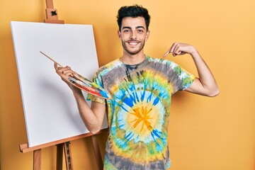 Young hispanic man standing drawing with palette by painter easel stand looking confident with smile on face, pointing oneself with fingers proud and happy.