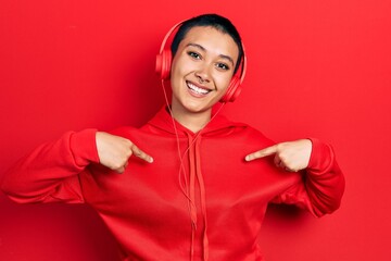 Beautiful hispanic woman with short hair listening to music using headphones looking confident with smile on face, pointing oneself with fingers proud and happy.