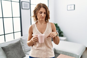 Sticker - Middle age woman drinking a cup coffee at home pointing down looking sad and upset, indicating direction with fingers, unhappy and depressed.