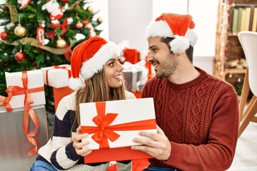 Canvas Print - Young hispanic couple wearing christmas hat opening gift at home.