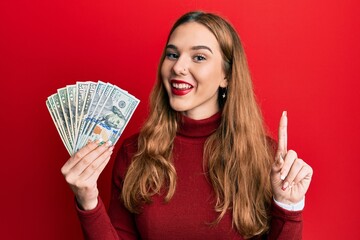 Sticker - Young blonde woman holding dollars smiling with an idea or question pointing finger with happy face, number one