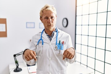 Poster - Young blond man wearing doctor uniform and stethoscope at clinic pointing down looking sad and upset, indicating direction with fingers, unhappy and depressed.