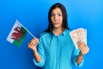 Sticker - Young latin woman holding wales flag and pounds banknotes skeptic and nervous, frowning upset because of problem. negative person.