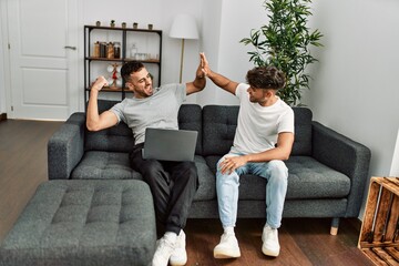 Wall Mural - Two hispanic men couple using laptop high five with hands raised up at home