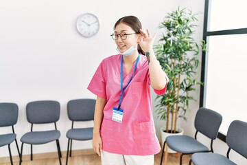 Sticker - Young asian nurse woman at medical waiting room smiling with hand over ear listening an hearing to rumor or gossip. deafness concept.