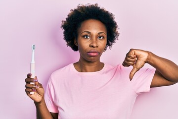 Wall Mural - Young african american woman holding electric toothbrush with angry face, negative sign showing dislike with thumbs down, rejection concept