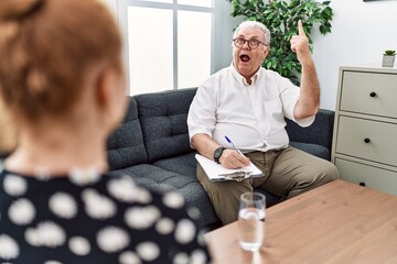 Wall Mural - Senior psychologist man at consultation office smiling amazed and surprised and pointing up with fingers and raised arms.