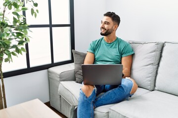 Sticker - Young handsome man with beard using computer laptop sitting on the sofa at home looking away to side with smile on face, natural expression. laughing confident.