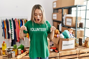 Sticker - Asian young woman wearing volunteer t shirt at donations stand pointing down with fingers showing advertisement, surprised face and open mouth